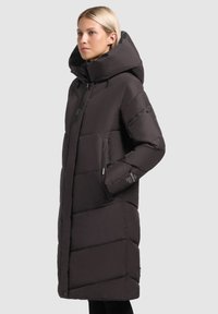 Kvinna i en lång svart vadderad kappa med stor huva, händer i fickorna, stående framför en enfärgad ljusgrå bakgrund.