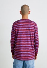 Långärmad skjorta i burgundy och lila randigt mönster, med rund halsringning och ribbad mudd. Tyget ser mjukt ut med en texturerad yta.