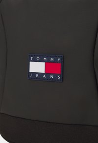 Zwarte stoffen rugzak met een rechthoekig logo-opdruk met "TOMMY" in het wit, "JEANS" in marineblauw en rood-witte accenten. Getextureerd oppervlak.