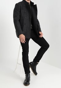 Zwart wollen mantel met dubbele rij knopen en een hoge kraag, gecombineerd met zwarte slim-fit broek en zwarte leren enkellaarsjes.