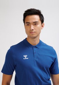 Niebieska koszulka polo z bawełnianej mieszanki, z kołnierzykiem na dwa guziki, krótkimi rękawami i białym wyszywanym logo na lewej piersi.