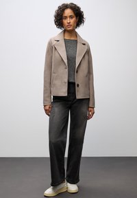 Beige skræddersyet frakke med reverskrave, sorte knapper, to sidelommer, båret over en grå sweater og sorte breddebenede jeans med hvide sneakers.