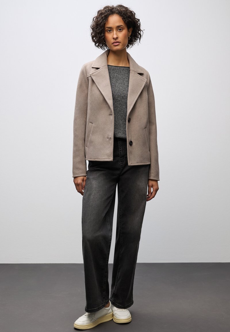 Cappotto beige su misura con colletto a rever, bottoni neri, due tasche laterali, indossato sopra un maglione grigio e jeans neri a gamba larga con sneakers bianche.