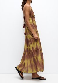 Robe longue avec un motif tie-dye jaune et marron, tissu fluide, bretelles ajustables et détails en tassels. Sandales plates noires portées.
