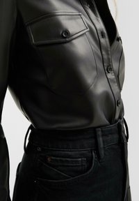 Zwarte leren shirt met een getailleerd ontwerp, voorzien van twee borstzakken en knoopsluitingen. Draagt met hoge taille zwarte jeans.