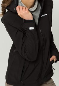 Giacca softshell nera con chiusura frontale a zip, cappuccio e tasche laterali. Presenta una texture liscia e dettagli del logo sul petto e sulla manica.