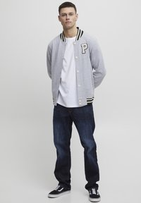 Grå varsity-jakke med svart og hvit stripet krage, hvit T-skjorte, mørkeblå jeans og svarte sneakers. Bokstaven "P" på jakken.