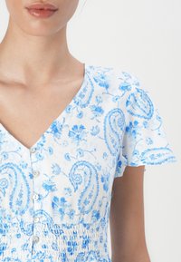 Robe à motif floral bleu et blanc avec un décolleté en V, des manches courtes et des boutons. Présente une taille froncée et un tissu doux et léger.
