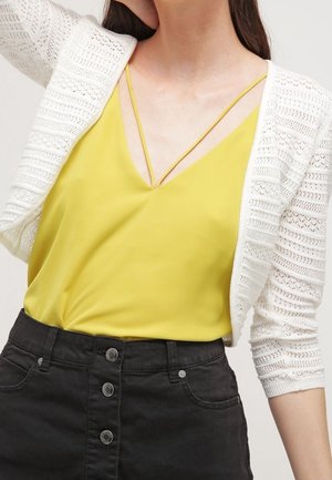 Femme portant un caraco jaune à fines bretelles sous un cardigan blanc en crochet et une jupe noire taille haute avec boutons sur le devant.