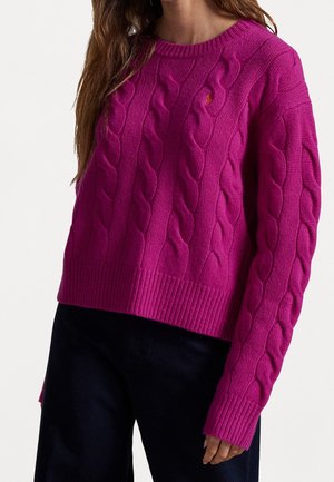 Femme portant un pull torsadé rose vif et un pantalon noir, vue recadrée montrant le haut du corps et la main.