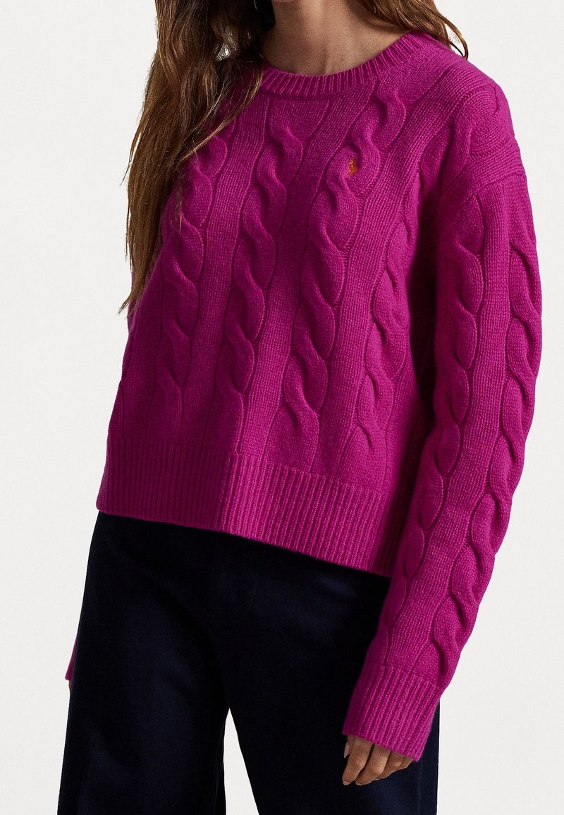 Femme portant un pull torsadé rose vif et un pantalon noir, vue recadrée montrant le haut du corps et la main.