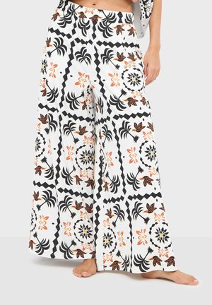 Białe spodnie o szerokich nogawkach z czarnymi i pomarańczowymi wzorami kwiatowymi i geometrycznymi, noszone boso przez stojącą osobę.
