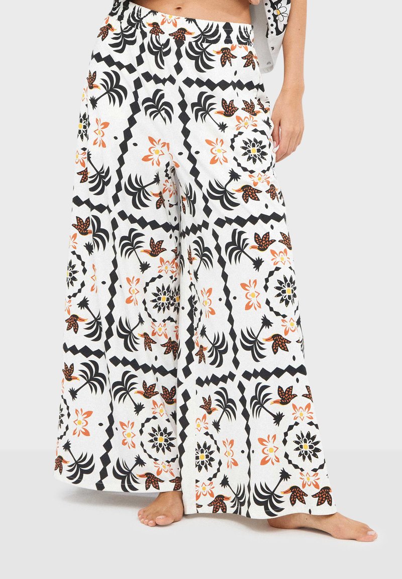 Pantalon blanc à jambes larges orné de motifs floraux et géométriques noirs et orange, porté pieds nus par une personne debout.