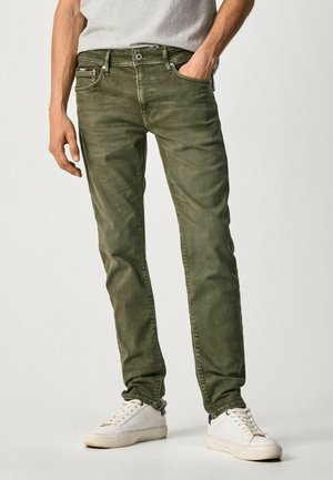 Mężczyzna noszący wyblakłe oliwkowo-zielone obcisłe dżinsy i białe buty sportowe, z jedną ręką w kieszeni i szarą koszulą wpuszczoną do środka.