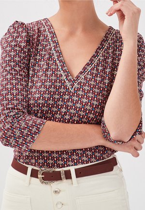 Femme portant un chemisier à motifs bordeaux avec un col en V, manches trois quarts, et un pantalon blanc, accessoirisée d'une ceinture marron avec une boucle décorative.