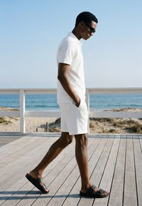 Homme portant une chemise blanche et un short avec des sandales noires marchant sur une terrasse en bois près de la plage sous un ciel bleu clair.