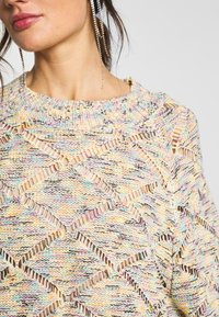 Pull en tricot avec un motif en losange et des découpes. Fil multicolore dans des tons pastel avec un col côtelé. Texture douce.