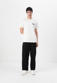 White cotton t-shirt featuring a small black graphic on the chest, paired with black trousers and white sneakers, set against a plain background.