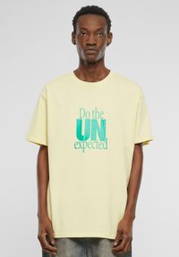Yellow cotton short-sleeve t-shirt with a crew neck. Front features turquoise text: "Do the UN expected." Relaxed fit, minimal design.