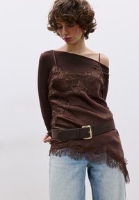 Hnedá čipkovaná košieľka vrstvená pod dlhým rukávom, so širokým čiernym opaskom so zlatou sponou, v kombinácii so svetlomodrými džínsami.