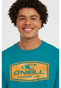 Camiseta de manga larga color verde azulado con un logo central de O'Neill en naranja, que presenta patrones de hojas tropicales y un neckline redondeado.