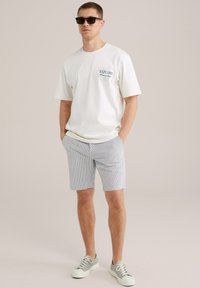Kortærmet hvid t-shirt med "EXPLORE" grafik, parret med mønstrede grå og hvide shorts og grå sneakers. Sorte solbriller bæres.