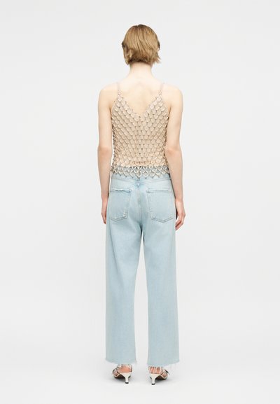 Άτομο με κοντά μαλλιά που φοράει ένα αμάνικο, ανοιχτού πλέγματος μπεζ τοπ, ανοιχτό μπλε τζιν ψηλόμεσο και λευκά σανδάλια με τακούνι, στέκεται με την πλάτη προς τα εμπρός.