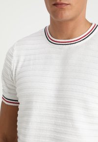Vit texturerad stickad skjorta med horisontella ränder, med en ribbad krage och ärmslut i röda och svarta detaljer.
