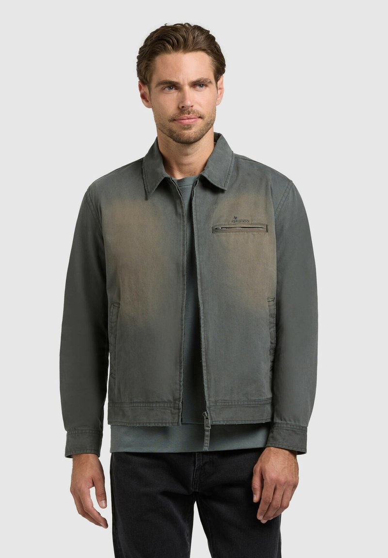 Férfi halványzöld, cipzáros kabátban, gallérral, kék inggel és fekete nadrágban, világosszürke háttér előtt állva.