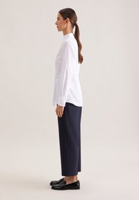 Hvid langærmet skjorte med mandarin-krave, kombineret med mørkeblå bukser og sorte loafers, vist i profil.