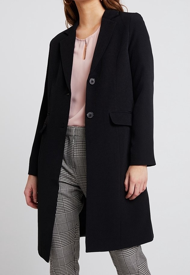 Femme portant un manteau noir cintré, un chemisier rose et un pantalon à carreaux noir et blanc, debout devant un fond clair uni.