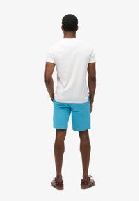 Camiseta blanca de manga corta, combinada con pantalones cortos azul brillante y sandalias de cuero marrón. Vista desde la parte posterior. Diseño sencillo y casual.