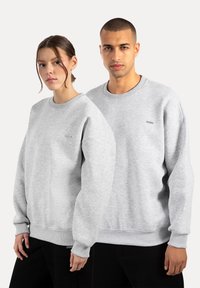 Grå sweatshirts lavet af blødt stof, med lange ærmer, ribstrikkede manchetter og et lille logo på brystet. Båret af to modeller.