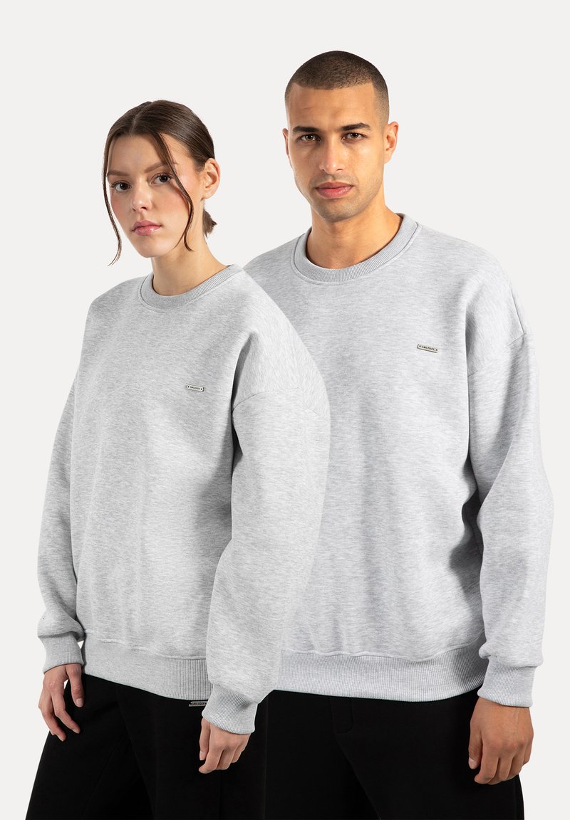 Grå sweatshirts lavet af blødt stof, med lange ærmer, ribstrikkede manchetter og et lille logo på brystet. Båret af to modeller.