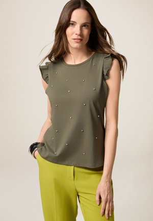Camicia verde oliva a maniche corte con spalle arricciate, decorata con piccole borchie argentate, abbinata a pantaloni verde brillante.