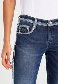 Jeans in denim di colore blu scuro, con orli sfilacciati, una vita strappata e una tasca con dettagli in rosso.
