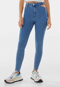 Vaqueros azules de mezclilla de tiro alto con detalles recortados en los costados, combinados con zapatillas deportivas multicolor con suelas gruesas.