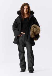 Cămașă neagră izolată cu glugă din blană sintetică, asociată cu pantaloni texturați închisi la culoare. Într-o mână ține o bucată de blană maro. Modelul stă în fața unui fundal simplu.