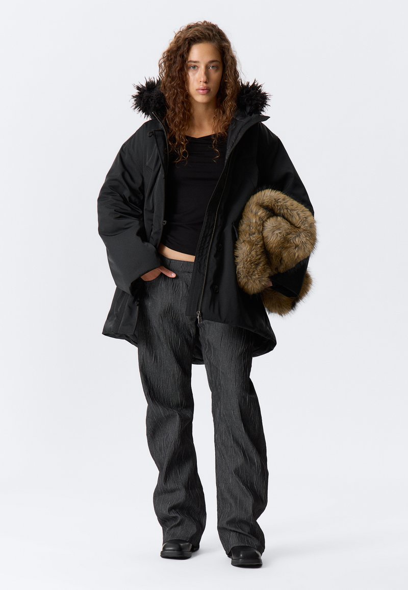 Cămașă neagră izolată cu glugă din blană sintetică, asociată cu pantaloni texturați închisi la culoare. Într-o mână ține o bucată de blană maro. Modelul stă în fața unui fundal simplu.
