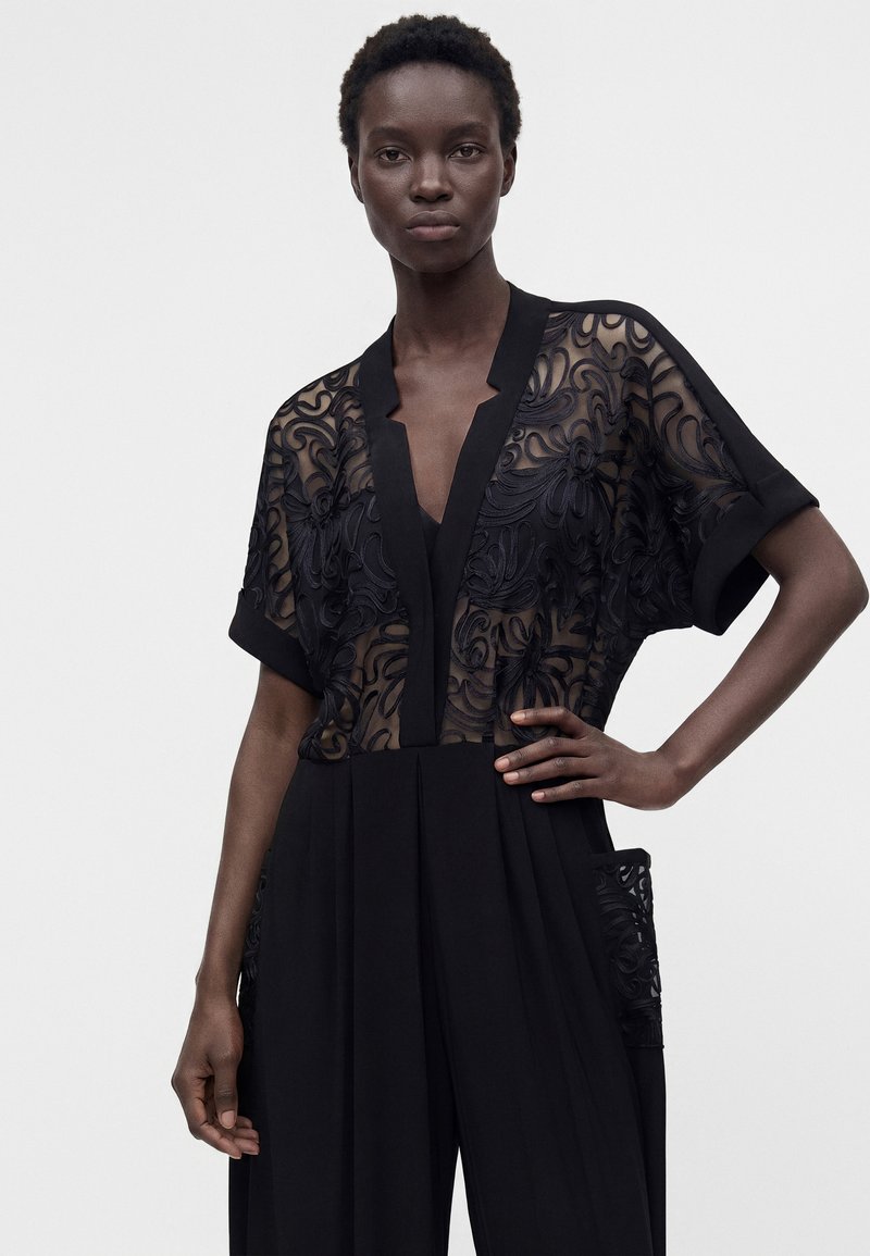 Femme aux cheveux courts portant un haut en dentelle noire transparente à motifs floraux et un pantalon noir, debout avec une main sur la hanche sur un fond uni.