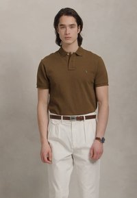 Noor mees seisab, kandes pruunist polo särk, valgeid plisseeritud pükse, pruuni vööd ja käekella, halli tausta ees.