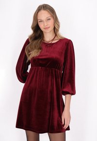 Rochie din catifea burgundă cu mâneci bufante, talie strânsă și detaliu sub formă de fundiță la decolteu. Textură netedă și design până la genunchi.