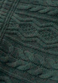 Dunkelgrüner gestrickter Stoff mit strukturierten Zopfmustern. Die Textur ist weich und geschichtet, mit subtilen Farbvariationen im gesamten Stoff.