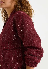 Chaqueta texturizada burdeos con un patrón tejido, mangas enrolladas y acentos metálicos. Incluye un detalle de bolsillo frontal.