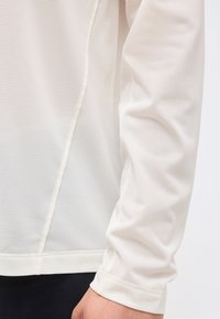 Κοντινό πλάνο ενός ατόμου που φοράει λευκό μπλουζάκι με μακριά μανίκια από δίχτυ με μικρές οπές και εμφανείς ραφές.