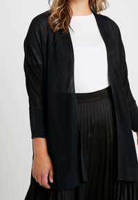 Černý, poloprůhledný kardigan s volným střihem, otevřeným předním dílem a třičtvrtinovými rukávy, doplněný bílou košilí a skládací černou sukní.