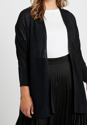 Zwarte, semi-doorzichtige cardigan met een losse pasvorm, open voorkant en driekwartmouwen, gecombineerd met een wit shirt en een geplooide zwarte rok.