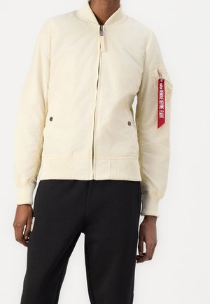 Person trägt eine cremefarbene Bomberjacke mit Fronttaschen und einem roten "Remove Before Flight"-Etikett am linken Ärmel, kombiniert mit schwarzen Hosen.