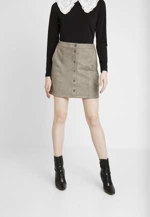 Femme portant une mini-jupe en suède taupe à boutons sur le devant, un haut noir à manches longues avec un col en dentelle blanche, et des bottines noires.