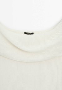 Pull à col roulé en maille côtelée blanche avec un col doux et replié, et une petite étiquette de marque noire cousue à l'intérieur de l'encolure.